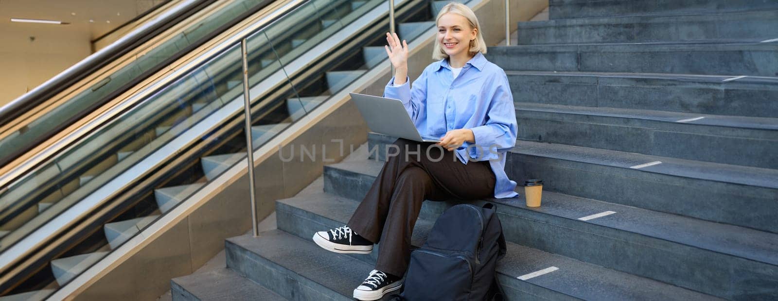 Girl student sitting on stairs in university campus, using laptop, working on project, doing homework, e-learning outdoors, drinking coffee outside, wearing stylish blue shirt.