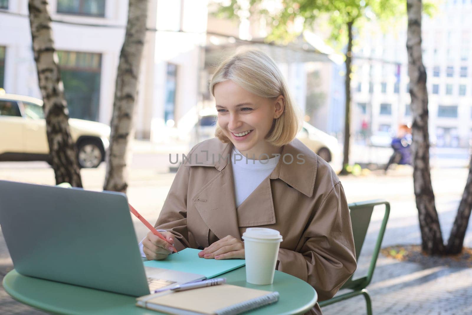 Young woman, freelancer, student doing homework in outdoor cafe, drinking her coffee on street, using laptop, connects to online lecture or doing course in internet by Benzoix