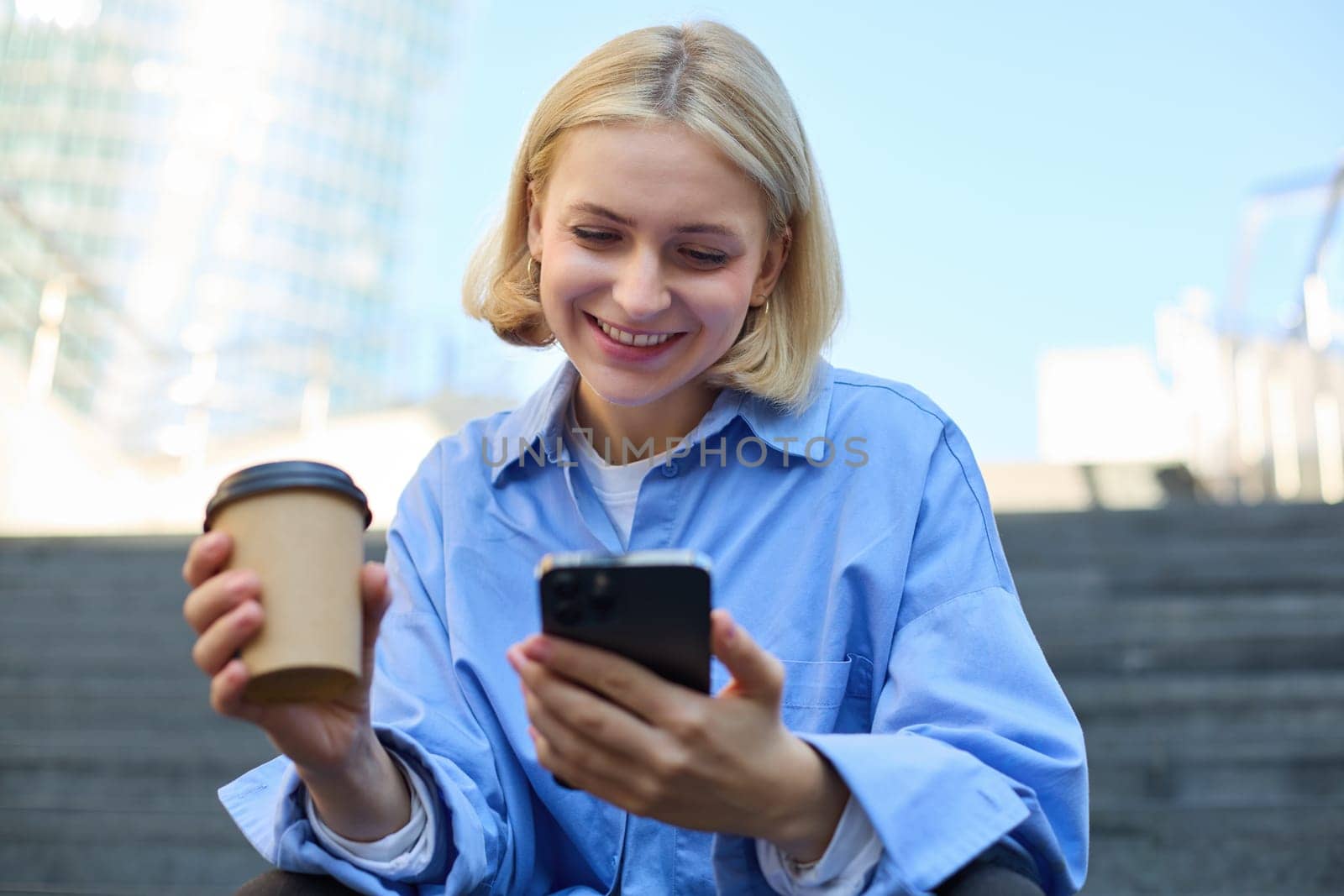 Close up portrait of young smiling woman with cup of coffee, drinking and sitting on stairs in city, holding smartphone in hand by Benzoix