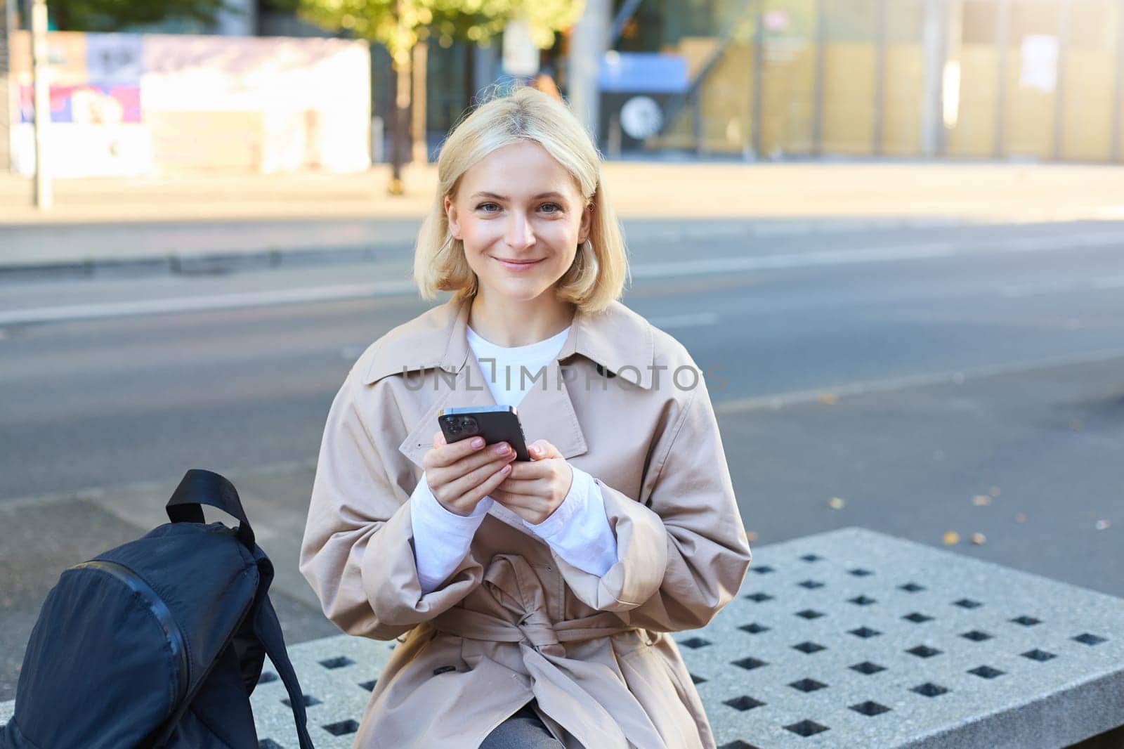 Outdoor shot of young blonde woman on street, sitting on bench with mobile phone, smiling at camera, waiting for friend outside of building, having a break by Benzoix