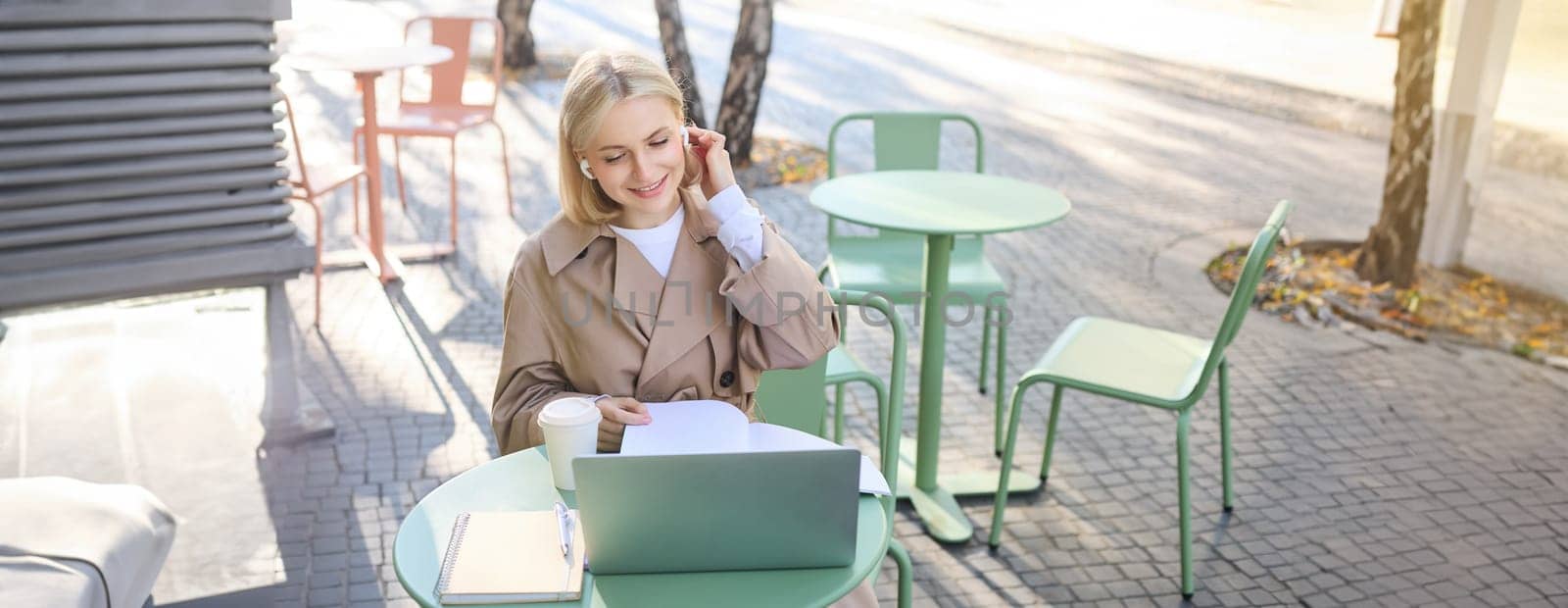Lifestyle and technology concept. Young blond working woman in trench coat, studying, working on project on laptop, sitting in coffee shop on sunny day, making notes, attending online course.