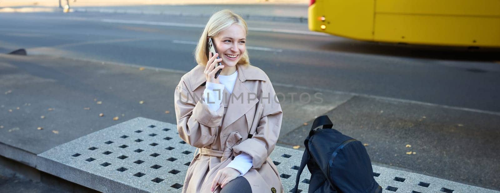 Lifestyle portrait of young beautiful blond girl, sitting on bench outside, talking on mobile phone, chatting with friend and laughing from funny conversation by Benzoix