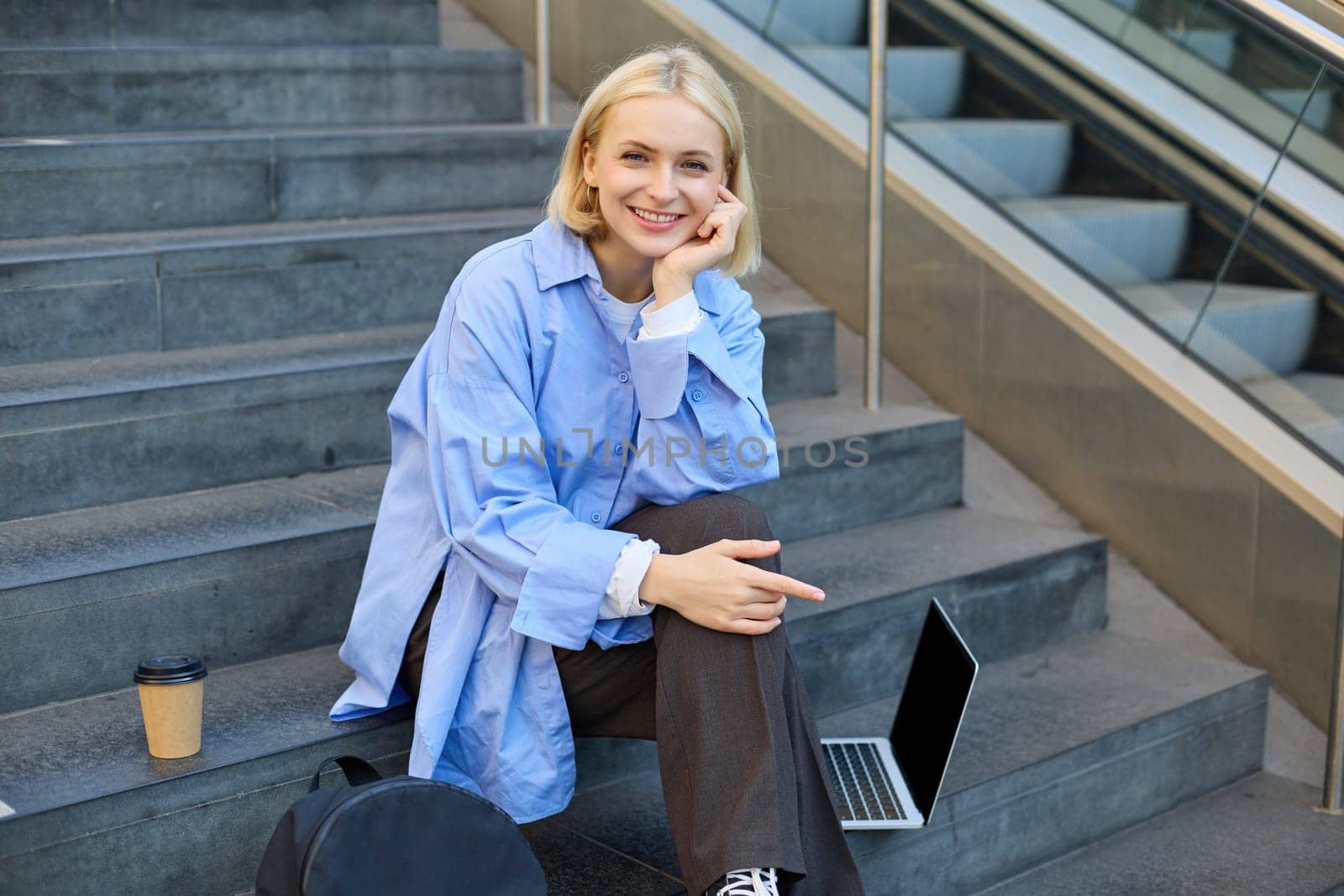 Portrait of young modern woman, student or freelancer, sitting on outdoor stairs, resting in city, has laptop and coffee, looking at camera with confident, smiling face.