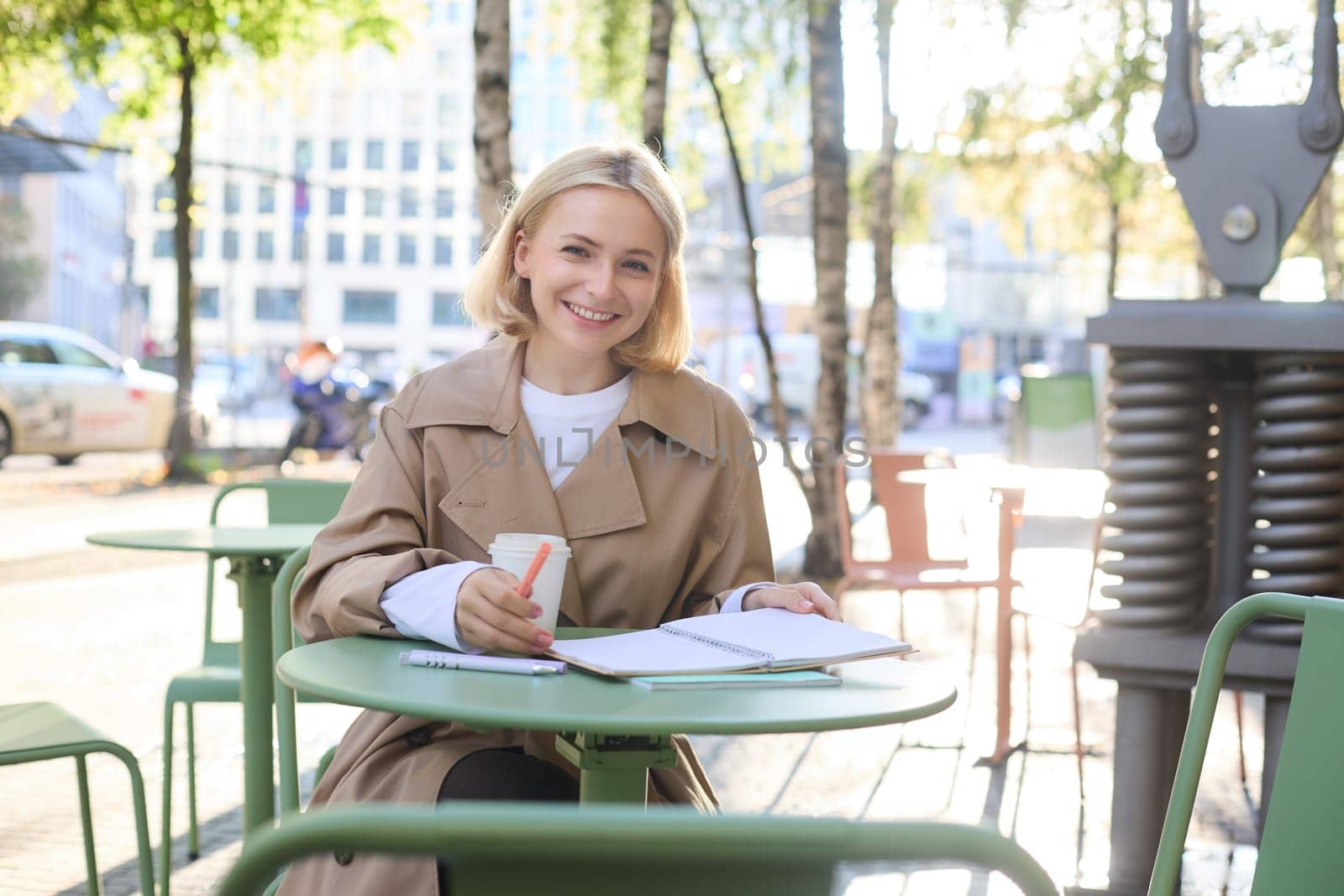 Portrait of young blonde woman sitting in cafe, doing homework and drinking coffee, writiing in journal, using notebook on fresh air by Benzoix