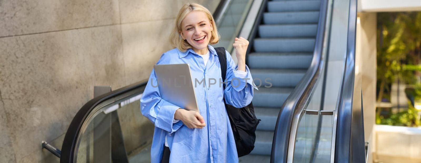 Portrait of enthusiastic young woman, makes fist pump, celebrating or triumphing, holding laptop and backpack, standing near escalator by Benzoix