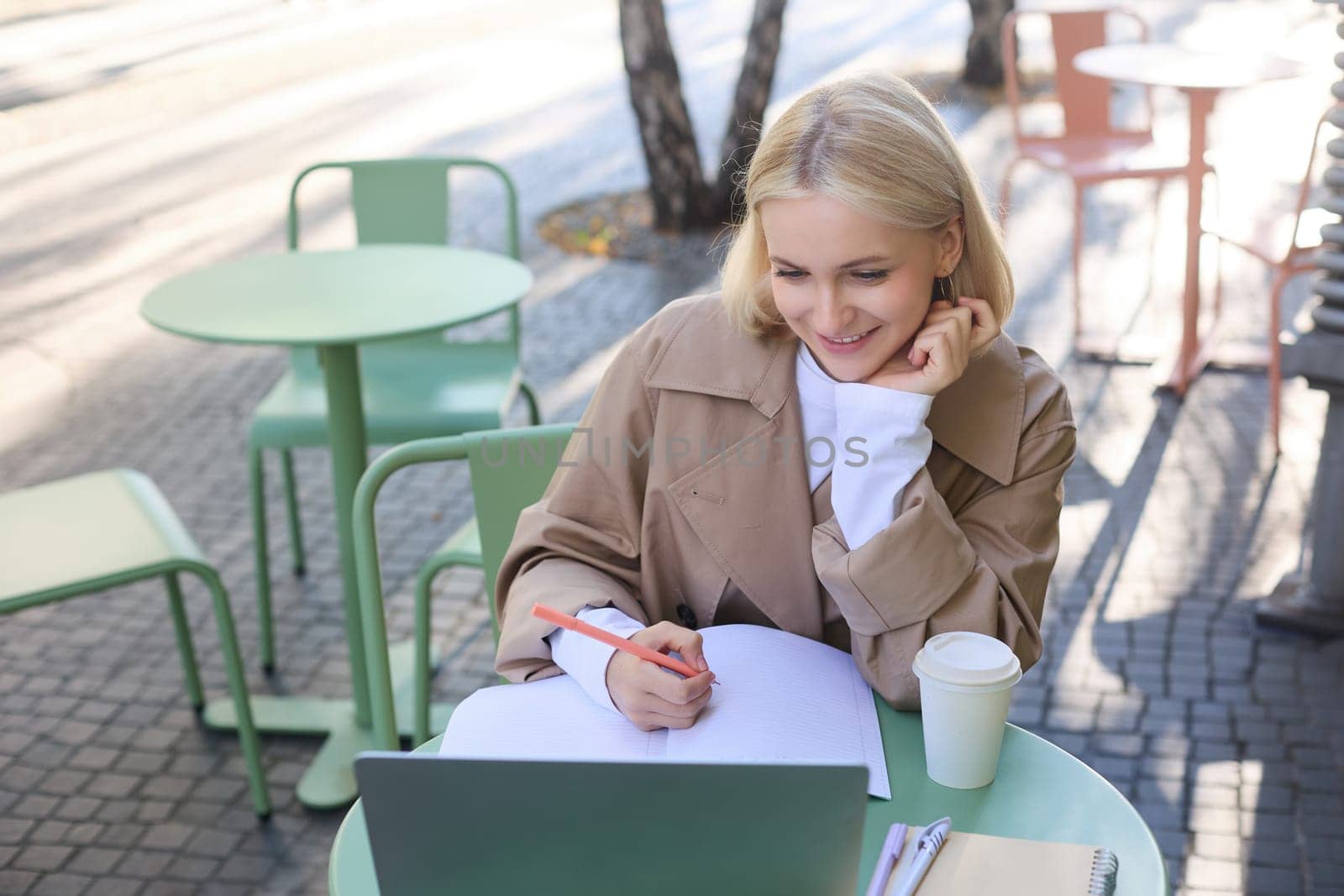 Image of smiling young female student, working on project, sitting outside on sunny day in cafe, looking at laptop and making notes, studying, doing homework outdoors.