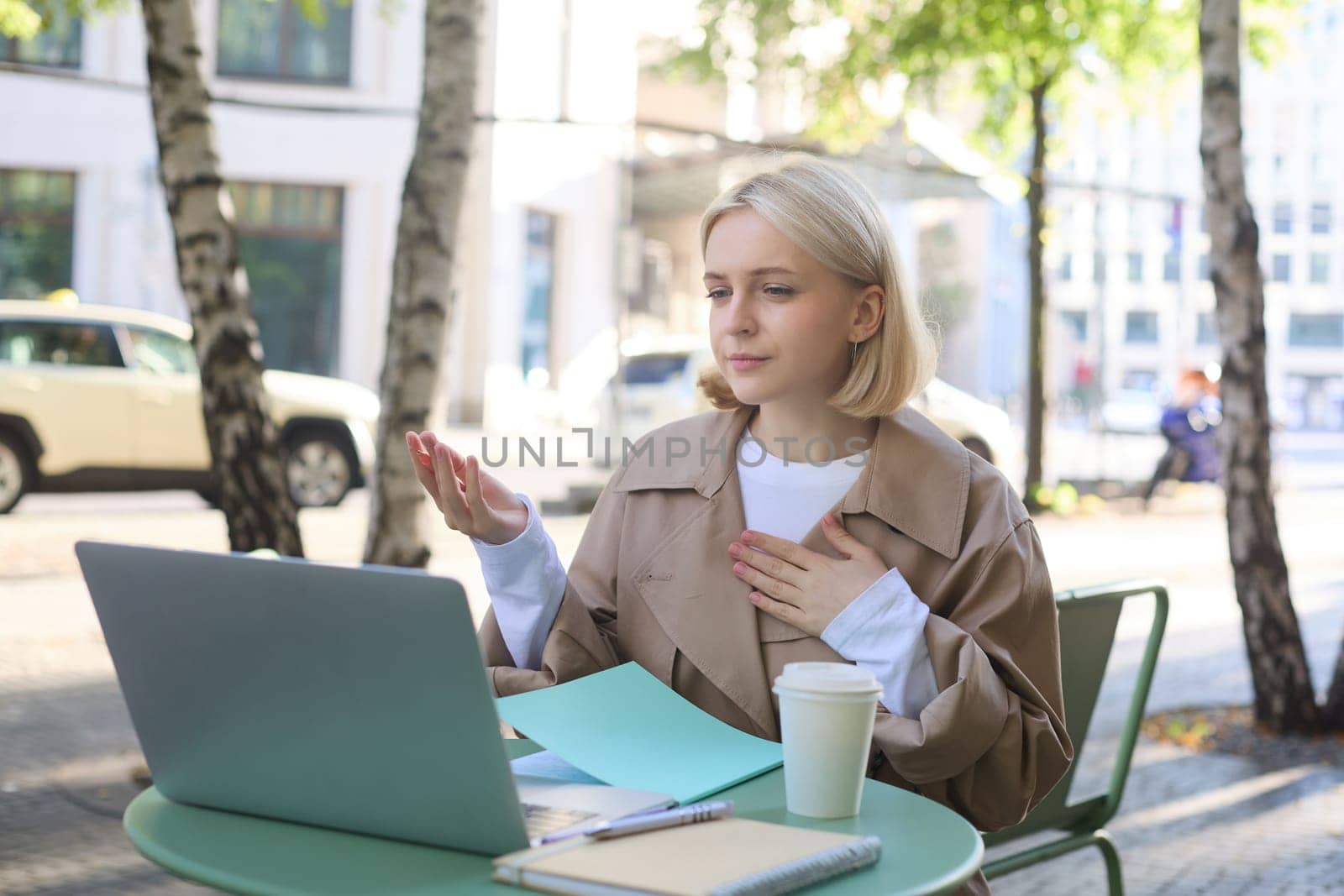Image of young stylish woman, sitting on street, drinking coffee in cafe, working remotely on laptop, attends online course or doing homework.