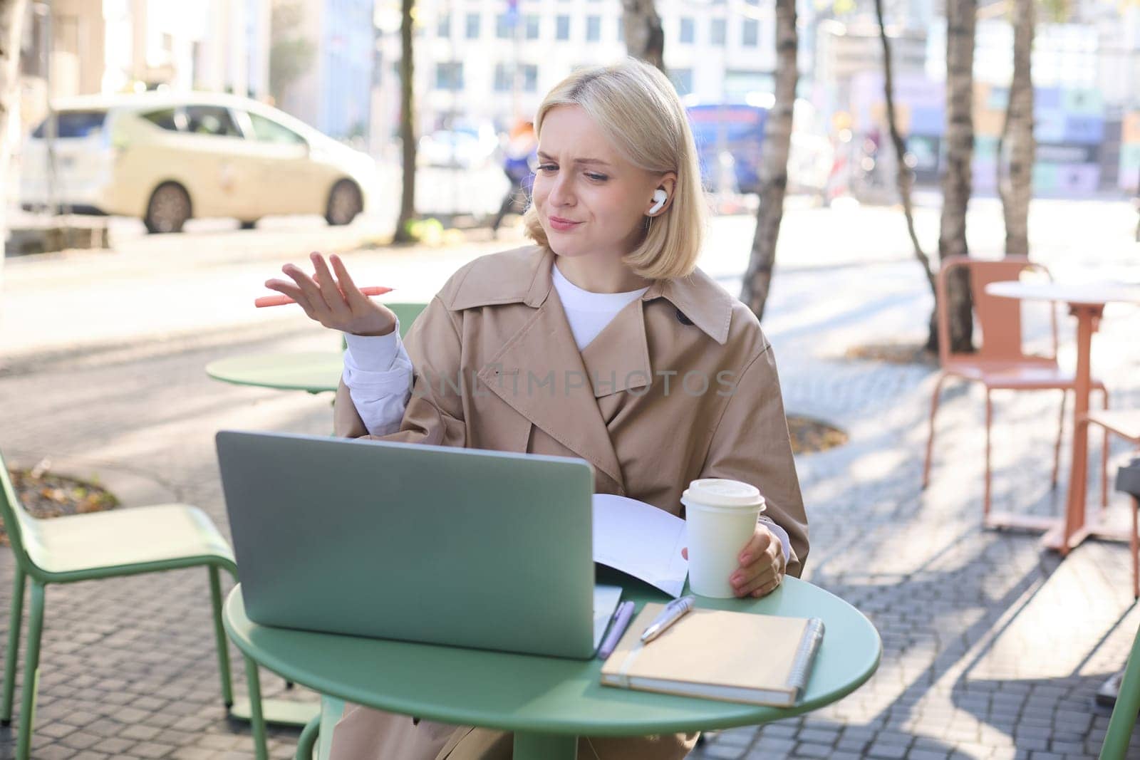 Image of young woman with puzzled face, looking at laptop in wireless headphones, confused while listening to lecture or online speaker, attend web course while sitting in outdoor cafe.