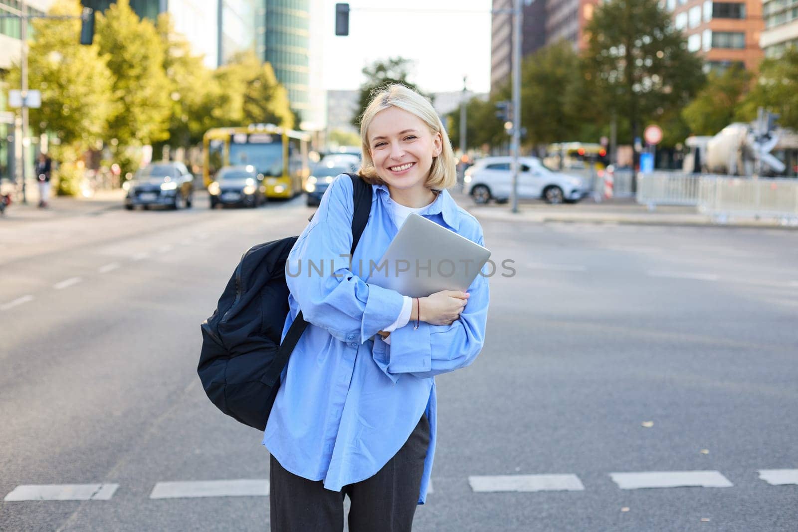 Image of young college student, happy female model on street, posing with backpack and laptop outdoors, with roads and cars behind her back, smiling at camera.
