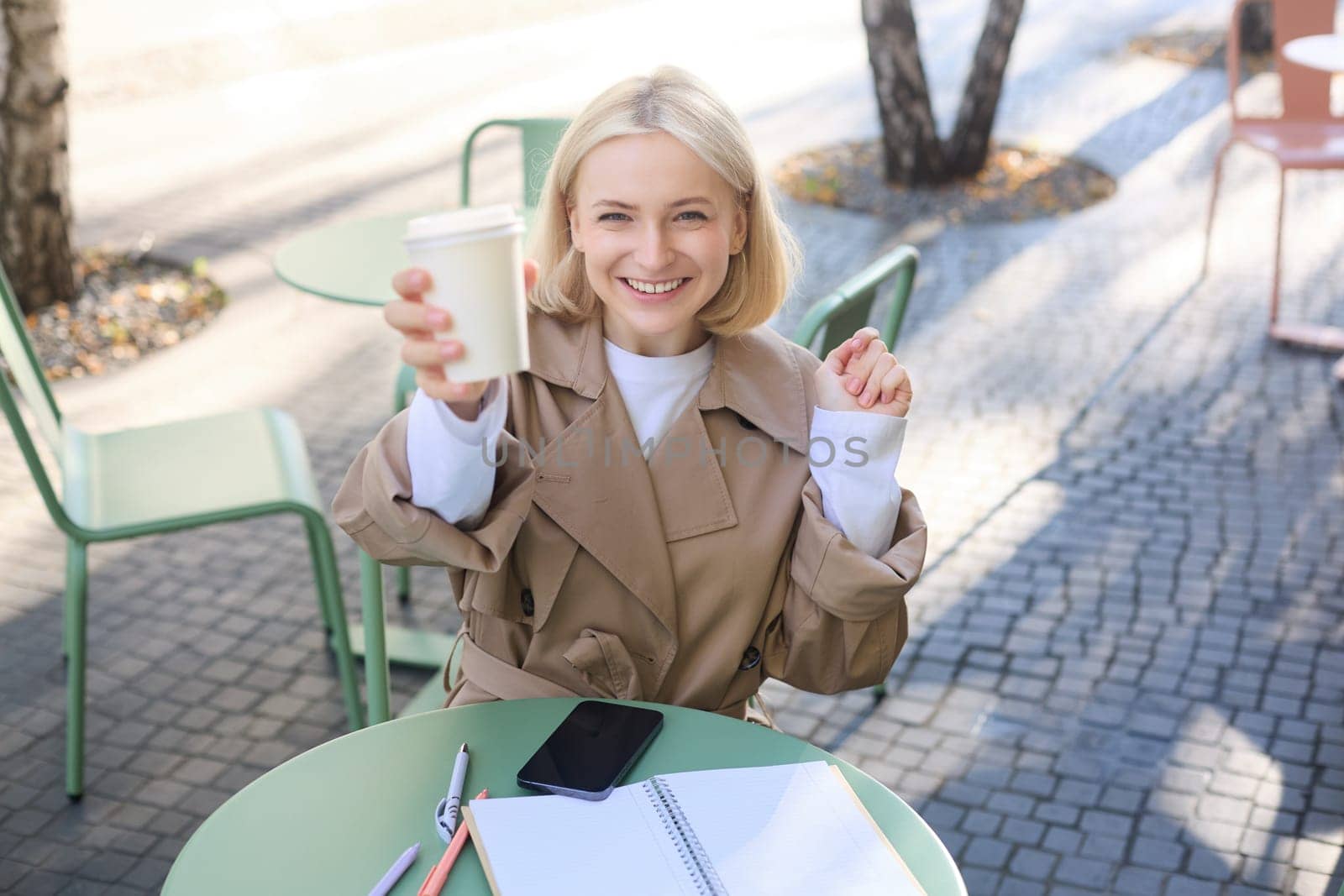 Beautiful blond woman, smiling, showing white takeaway coffee cup, drinking beverage in cafe shop, sitting outdoors.