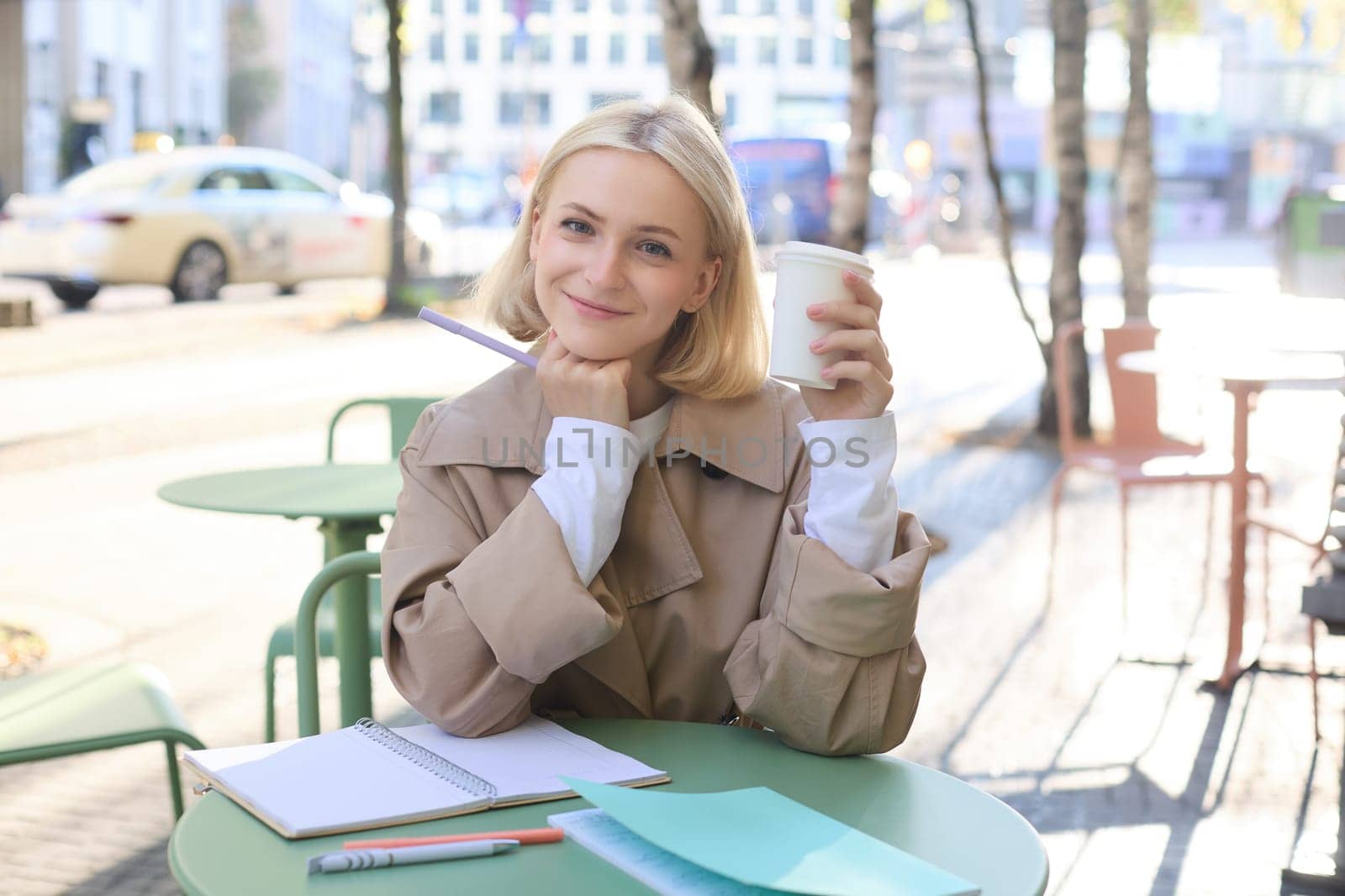 Stylish young urban woman, sitting in cafe and drinking coffee, smiling and looking at camera, doing homework or working on project, making notes in notebook.