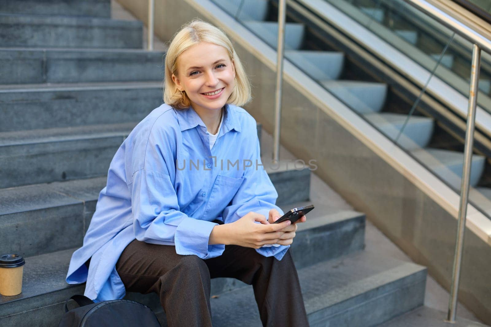 Lifestyle portrait of young urban female model, student drinks cup of takeaway coffee, sitting on stairs outdoors, using mobile phone, smiling and looking happy while taking a break.
