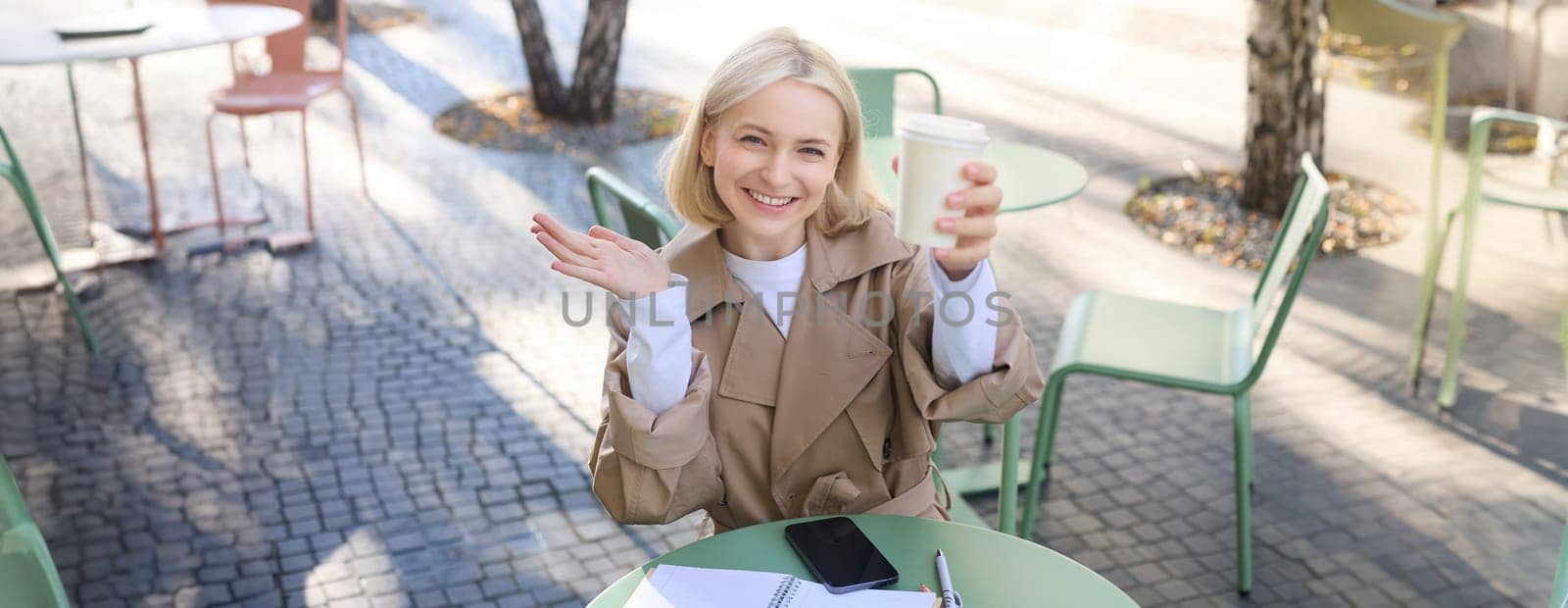 Food and drink concept. Young happy woman sitting in cafe, enjoying bright day outdoors, raising cup of coffee and smiling, drinking chai by Benzoix