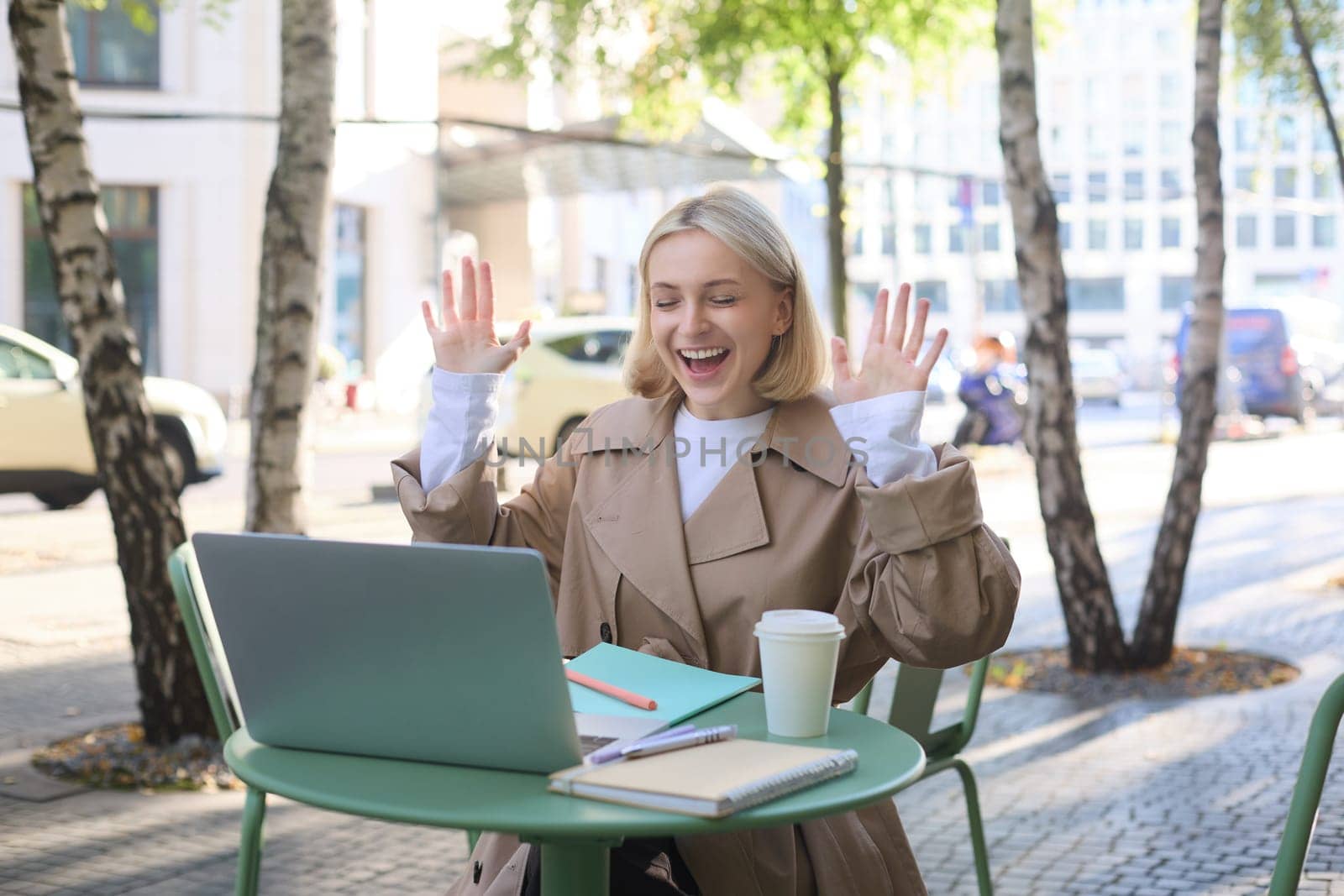 Enthusiastic young woman, carefree girl celebrating, sitting in outdoor cafe with laptop, raising hands up and laughing, triumphing.
