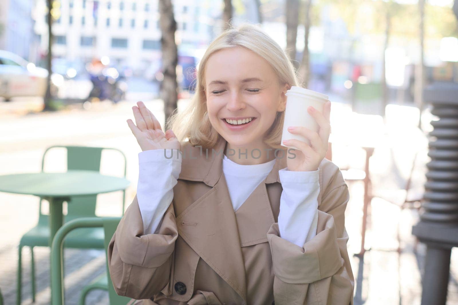 Portrait of cheerful young blond woman, enjoying spending time in outdoor cafe, drinking coffee and laughing, having casual meet up in street restaurant. Lifestyle concept