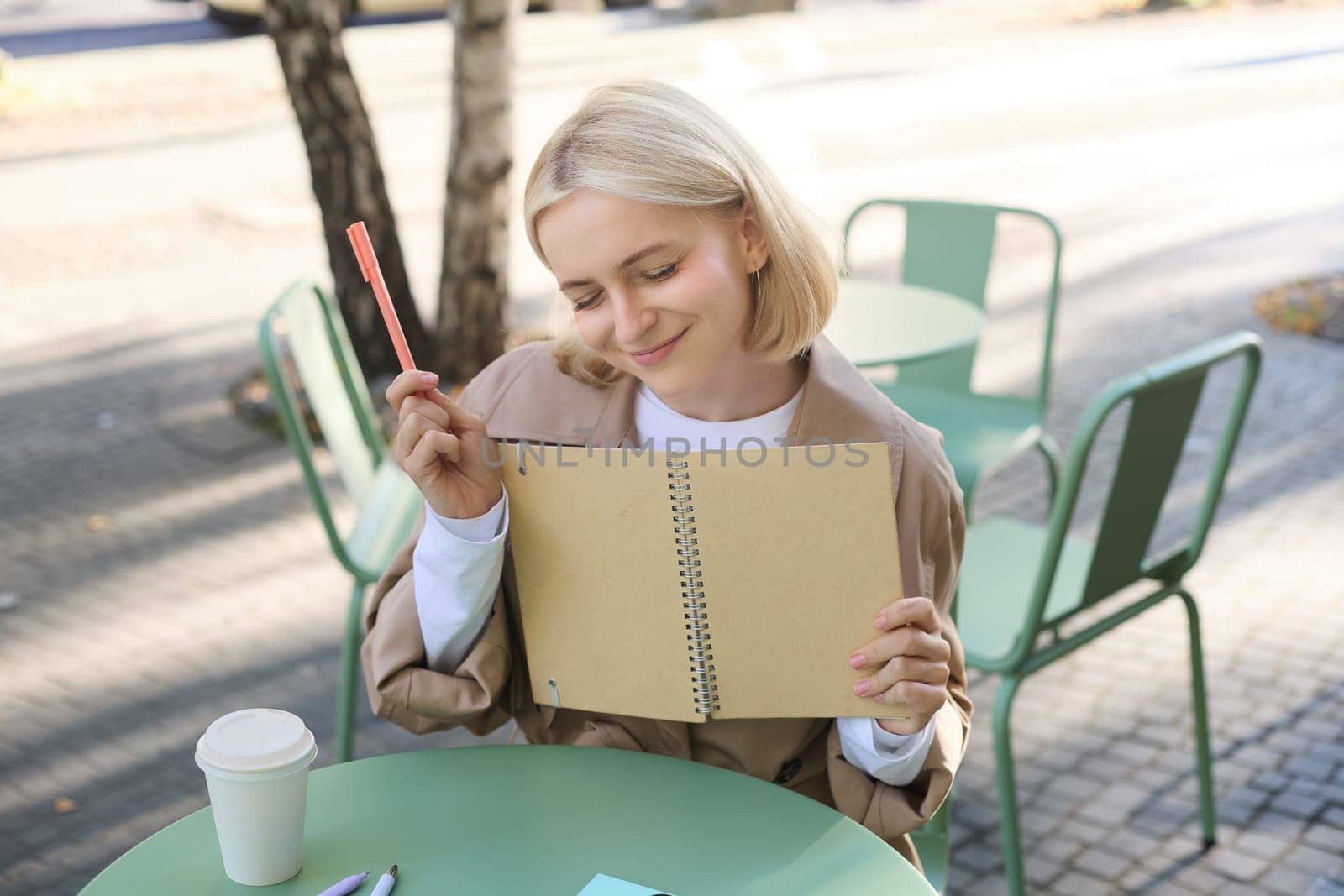 Portrait of young creative woman, doing sketches outdoors, sitting in coffee shop with notebook and pen, drawing art and smiling.