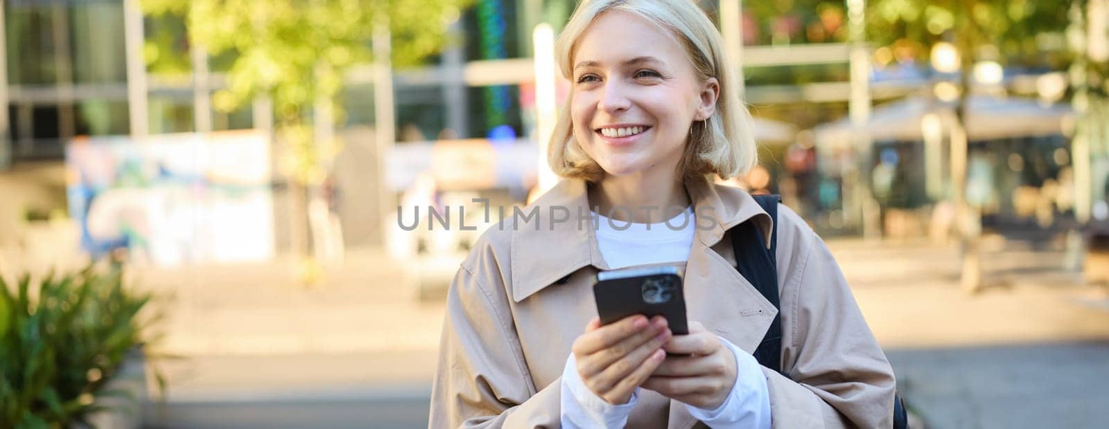 Carefree smiling woman standing on sunny street, using mobile phone, waiting for car or bus with smartphone, looking around by Benzoix