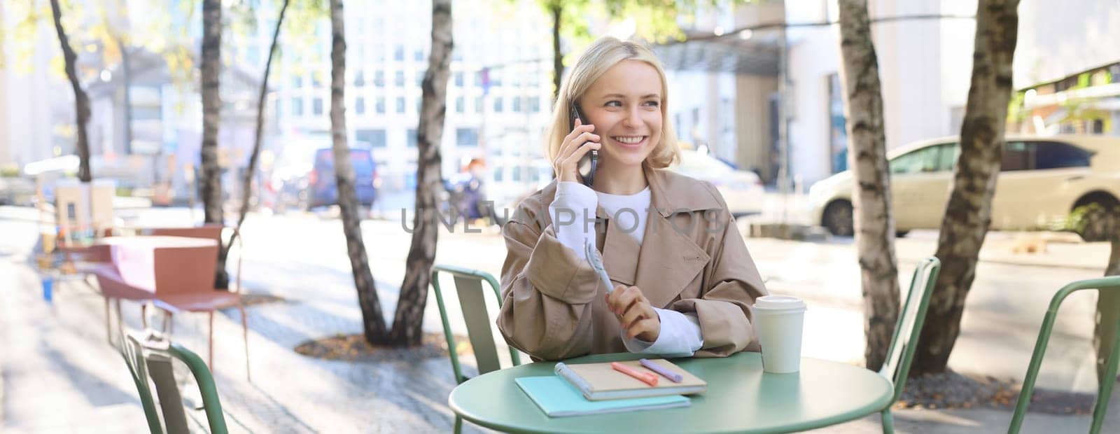Image of young modern female model in trench, sitting in cafe outdoors, has journals and notebooks on table, talking on mobile phone, answers a call and smiles by Benzoix