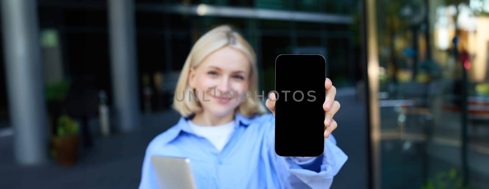 Portrait of beautiful young woman, student posing on street near campus, holding laptop, showing mobile phone screen, demonstrating app or promo on smartphone and smiling by Benzoix