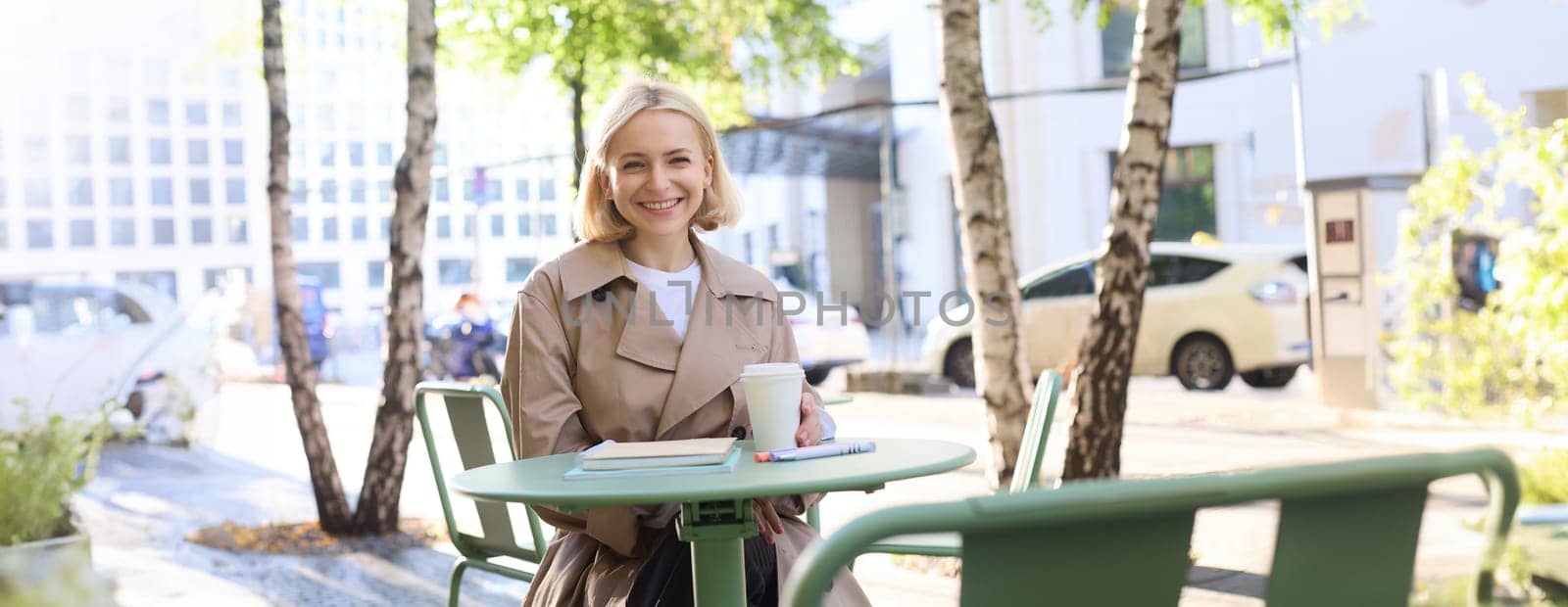 Urban lifestyle concept. Young beautiful woman sitting in outdoor cafe, contemplating city life, drinking coffee and smiling on street by Benzoix