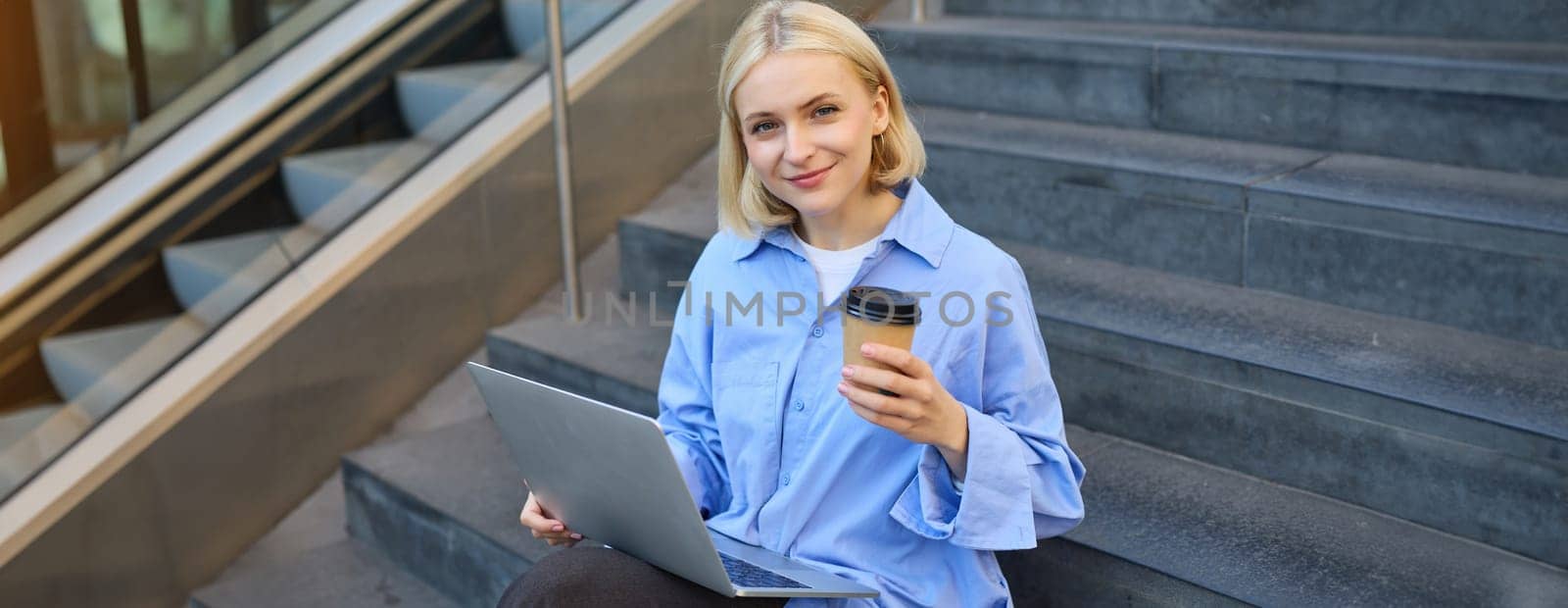 Close up portrait of stylish young female student, sitting on street stairs with laptop computer, holding coffee drink and smiling at camera.
