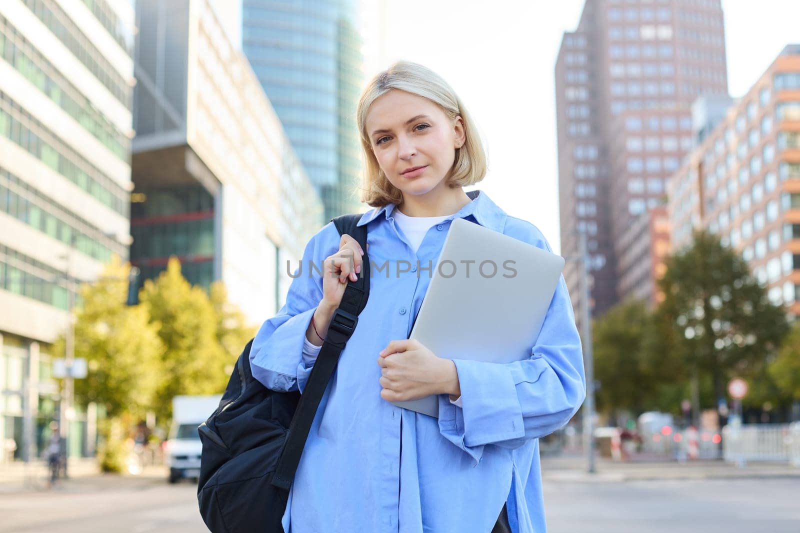 Portrait of young confident woman, college student with backpack and laptop, heading to lesson, standing outdoors on empty street with big buildings behind.