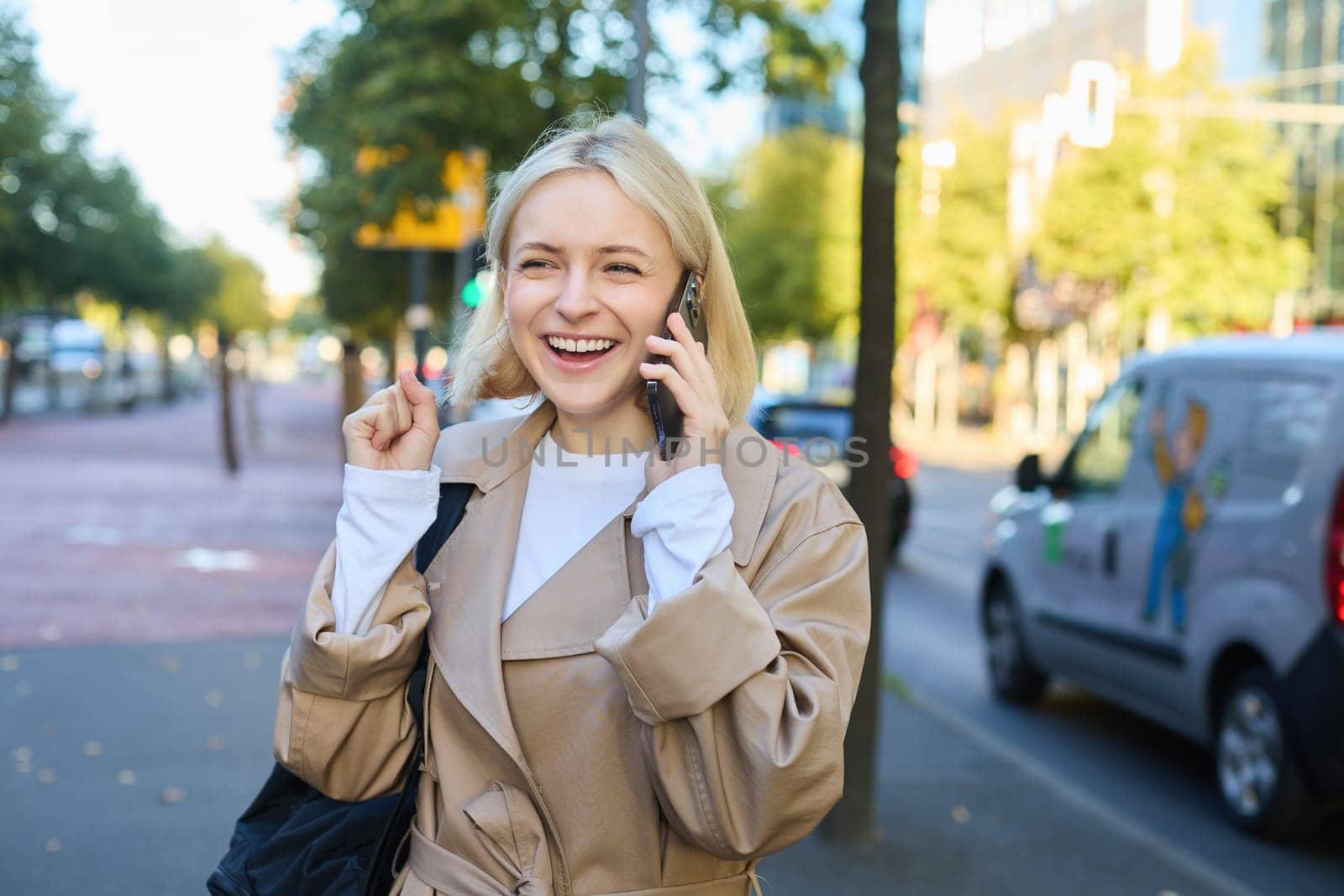 Portrait of cheerful girl talking on mobile phone, laughing while walking along the street, has backpack on shoulder by Benzoix