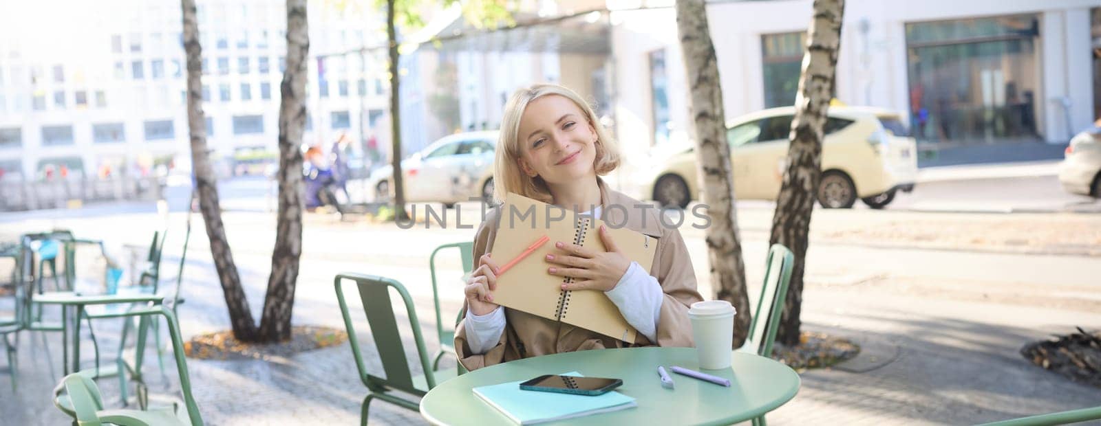 Image of young smiling blond woman sitting in an outdoor cafe, holding notebook, doing her homework outside in coffee shop, looking happy.