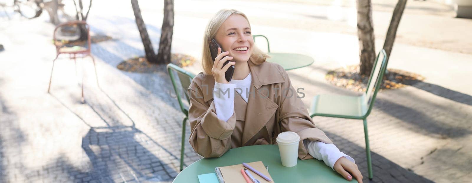 Cheerful young woman laughing, smiling while talking on mobile phone, sitting in cafe outdoors, enjoying friendly conversation.