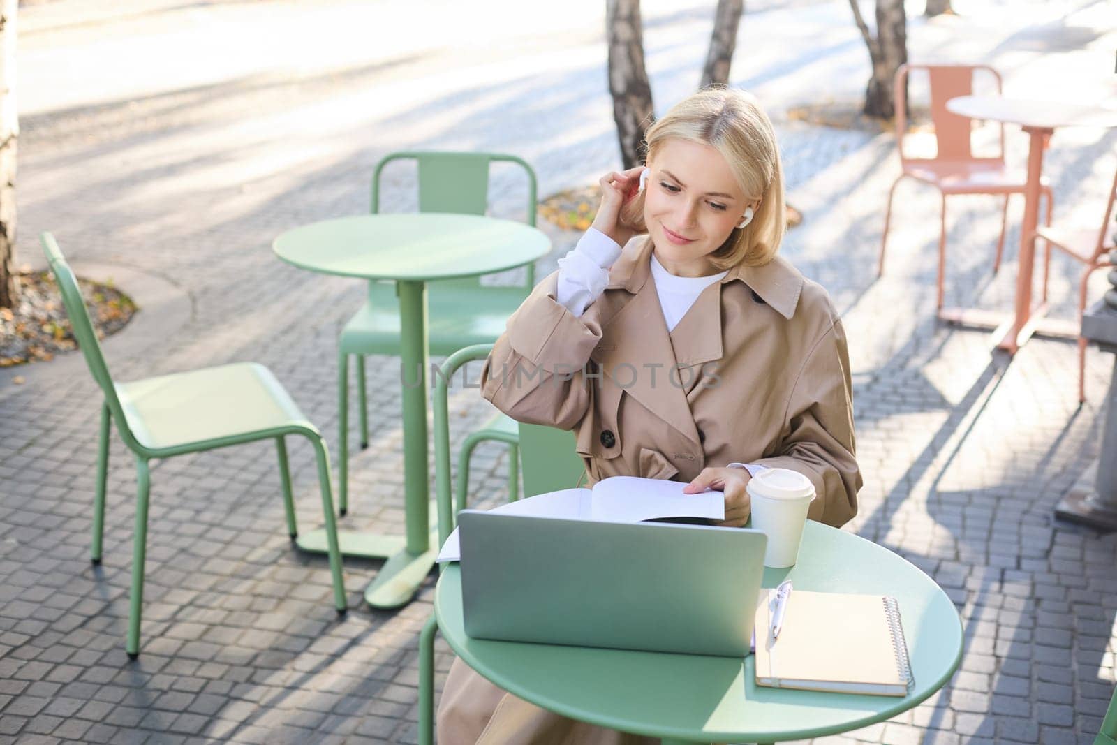 Lifestyle and people concept. Woman in outdoor cafe, sitting with laptop on street, connects to online course or lecture, doing homework, making notes, wearing wireless headphones, drinking coffee by Benzoix