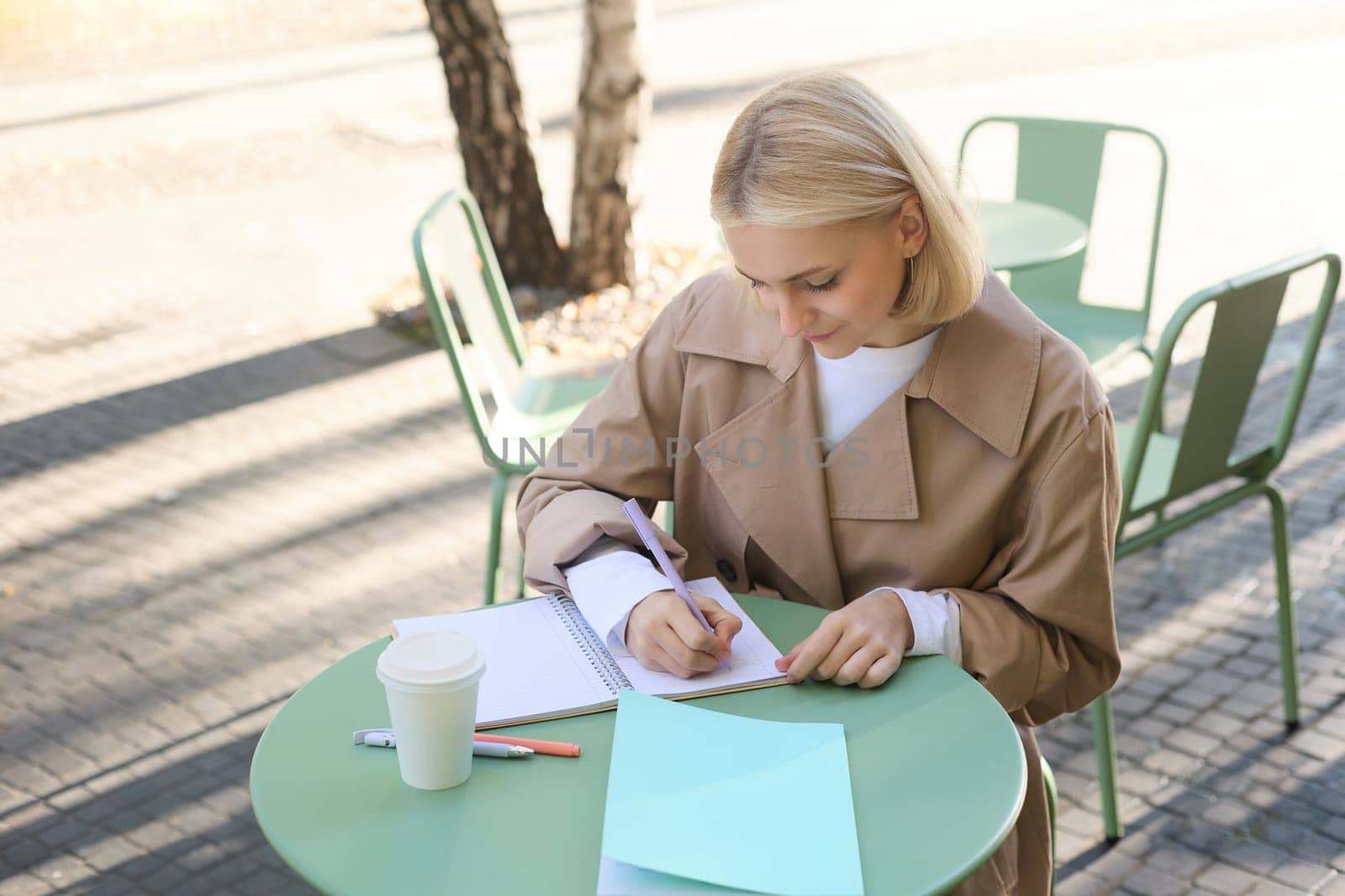 Portrait of woman working on project, writing in notebook, sitting outdoors in cafe, drinking coffee and doing homework by Benzoix