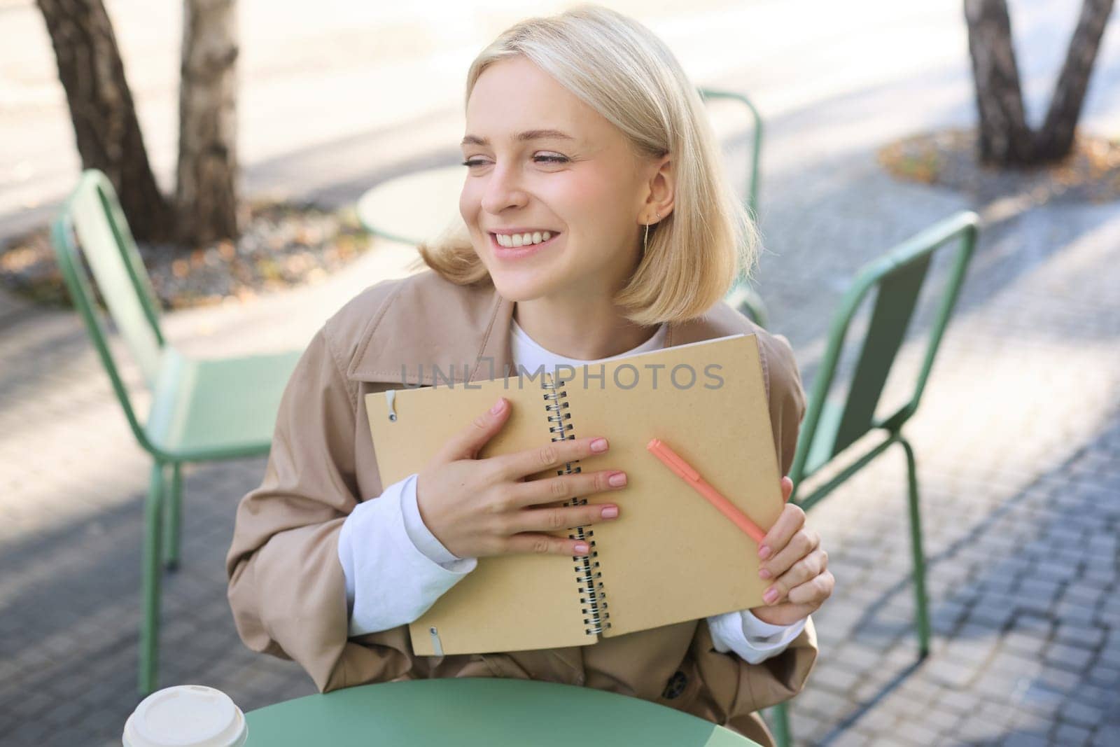 Portrait of carefree, smiling young woman with notebook and pen, sitting in outdoor cafe near table, writing in journal, making plans in her planner by Benzoix