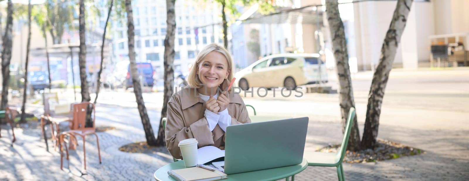 Portrait of excited blond woman, student in outdoor cafe, sitting in coffee shop with laptop, looking happy and amused, holding pen with notebook, studying or doing homework by Benzoix