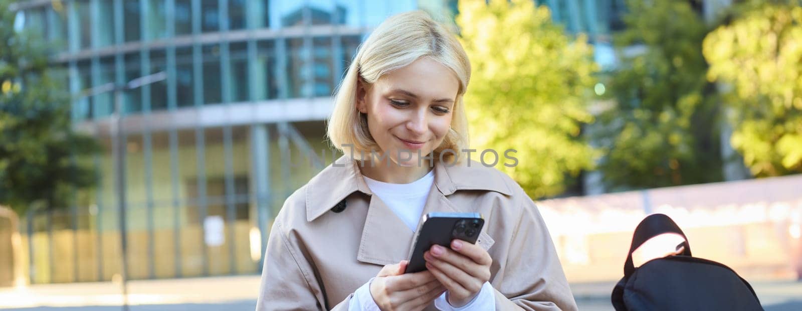 Close up portrait of smiling female model on street, sitting on bench with mobile phone, looking at smartphone screen, messaging with friend while waiting outside.