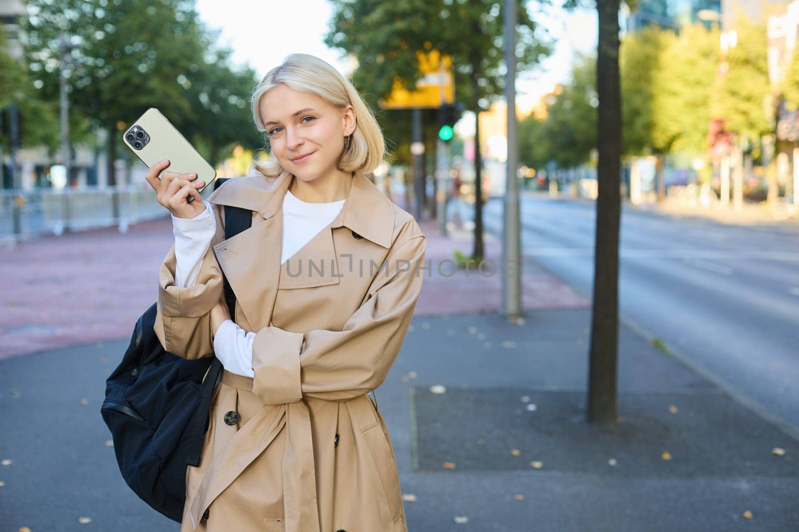 Image of young modern woman in trench coat, student holding mobile phone and backpack, posing on streets, standing near road, smiling and looking at camera by Benzoix