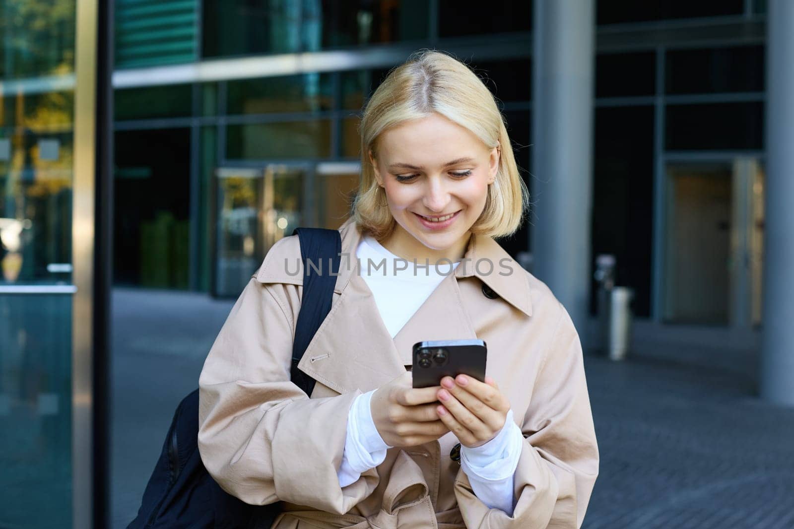 Portrait of young stylish woman walking in city centre with smartphone, using mobile phone app outdoors, waiting for someone, going to work by Benzoix