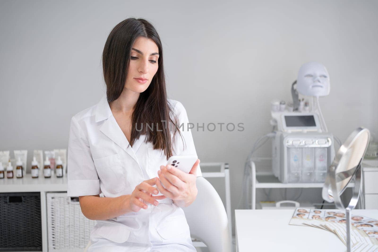 Cosmetologist in beauty salon sitting looking on phone screen. Dermatologist in beauty clinic plain facial skin treatments for clients in beauty salon using smartphone