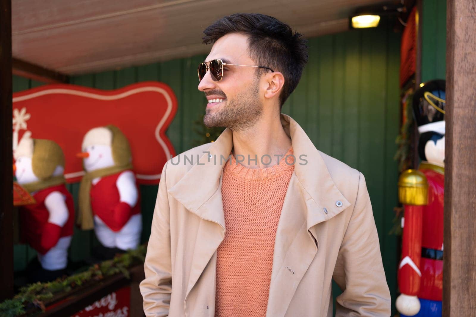 Stylish man in sunglasses smiles looking side standing outdoor. Bearded male standing near Christmas decoration outside. Happy people positive emotions