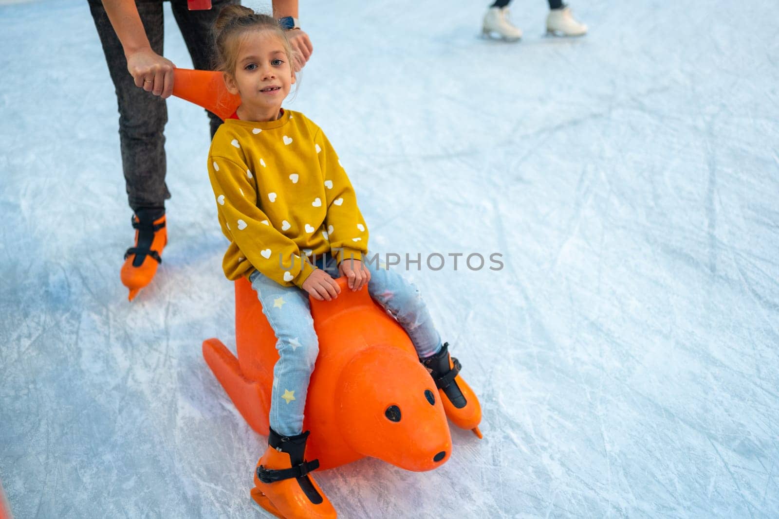 Child glides across ice rink on toy. Child glides across ice rink. Family socializing in winter, playing sports and having fun in snow. Daughter enjoying winter activities