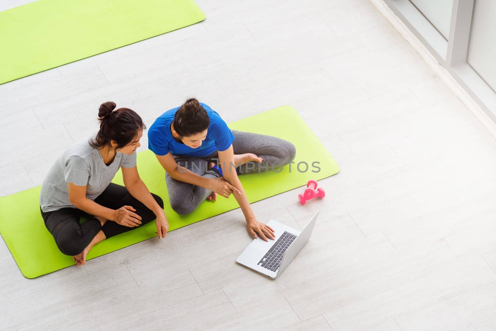 Overhead two Asian lifestyle adult and young woman in sportswear sitting floor on mat with dumbbells look laptop computer for training videos online in yoga studio, fit sport healthy workout exercise