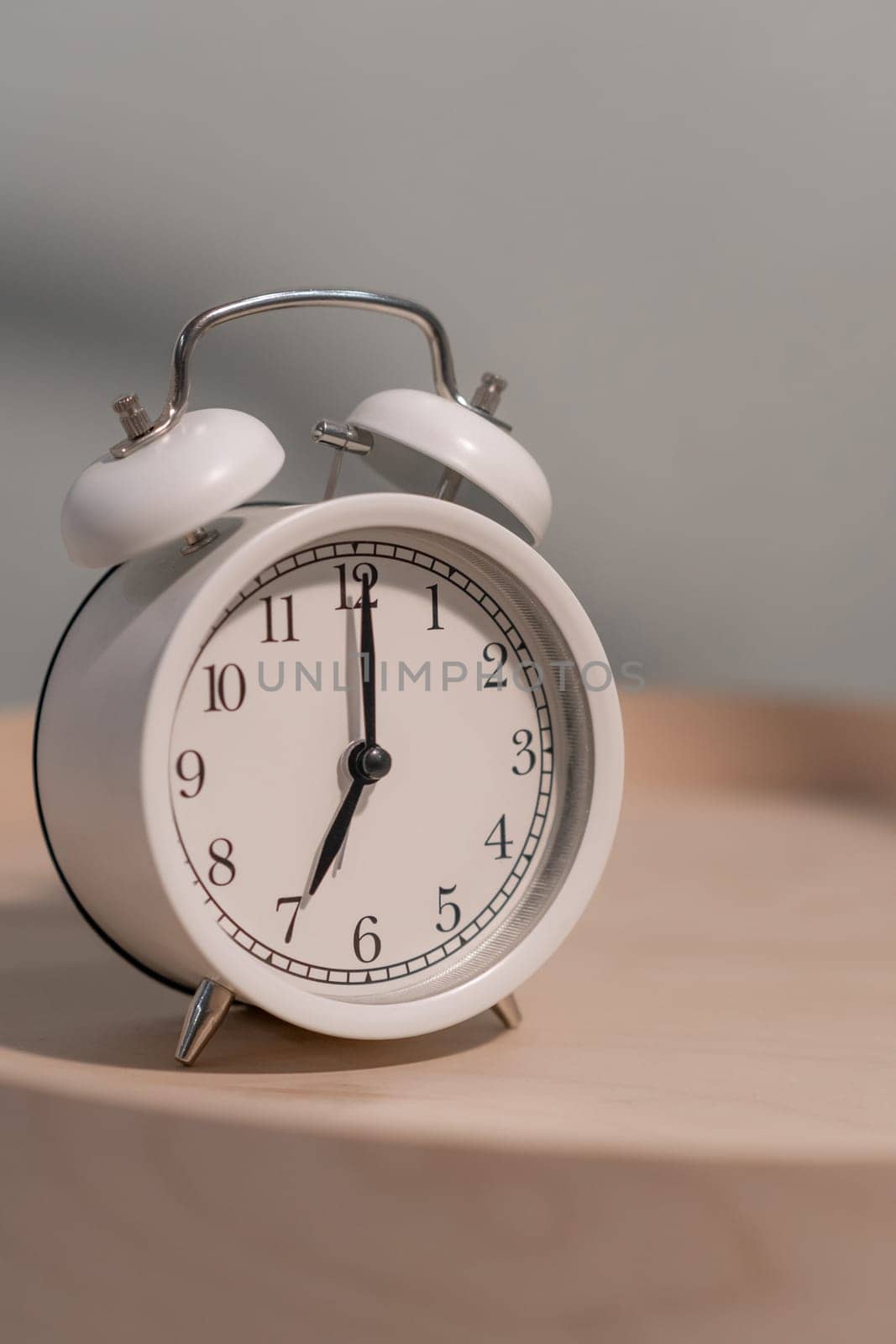 Close-up of a round white alarm clock on a table in the bedroom. The hands on the clock show seven o'clock in the morning, time to get up. Retro alarm clock on the table, vintage tone. space for text