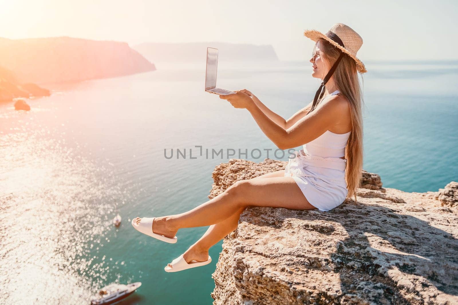 Working remotely on seashore. Happy successful woman female freelancer in straw hat working on laptop by the sea at sunset. Freelance, remote work on vacation