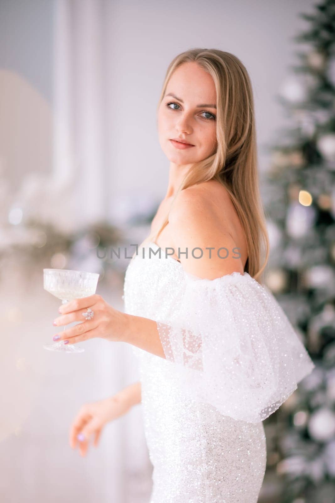 The blonde in the Christmas room. A beautiful blonde woman in a shiny light short dress with a train stands in a beautiful bright room decorated with a festive interior with a Christmas tree