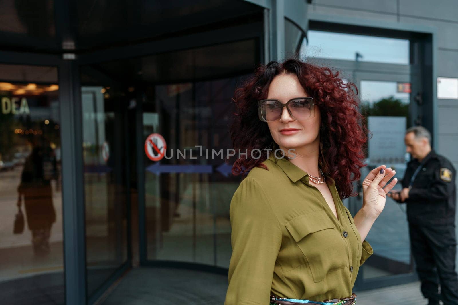 Woman leaves a supermarket. Caucasian model with long brunette hair, wears sunglasses and a khaki dress.