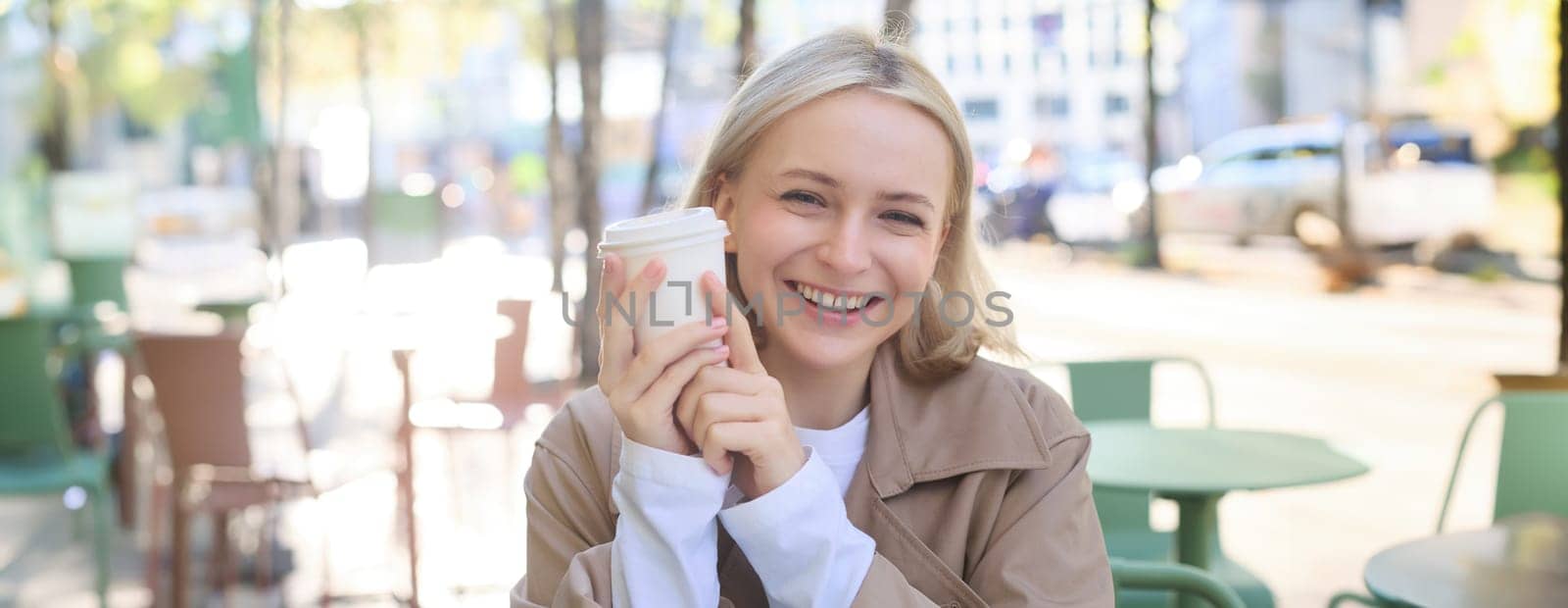 Close up portrait of smiling blonde woman, university student, holding cup of coffee, warming up her hands on chilly autumn day, sitting in outdoor cafe, laughing at camera.