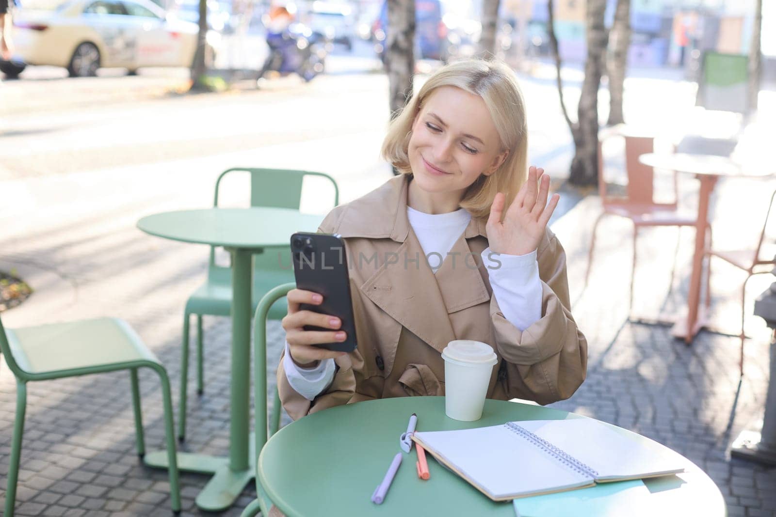 Portrait of young stylish woman in trench, sitting outdoors in city cafe, talking on video chat, using mobile phone to connect to online conversation, drinking coffee.