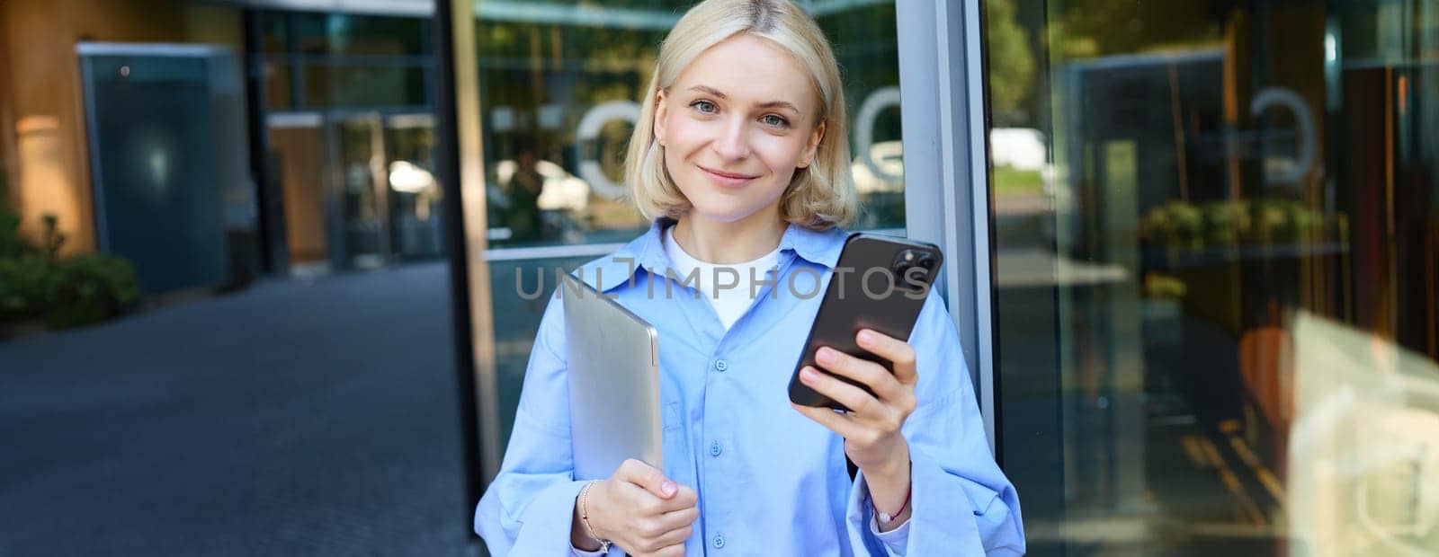 Lifestyle shot of young smiling woman, student or employee near office building, holding mobile phone and laptop in hands, looking happy, checking notification on smartphone.