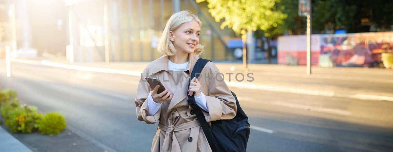Portrait of young blonde woman with smartphone, holds backpack, waits for taxi on street, looking at the road and smiling, using mobile phone app.