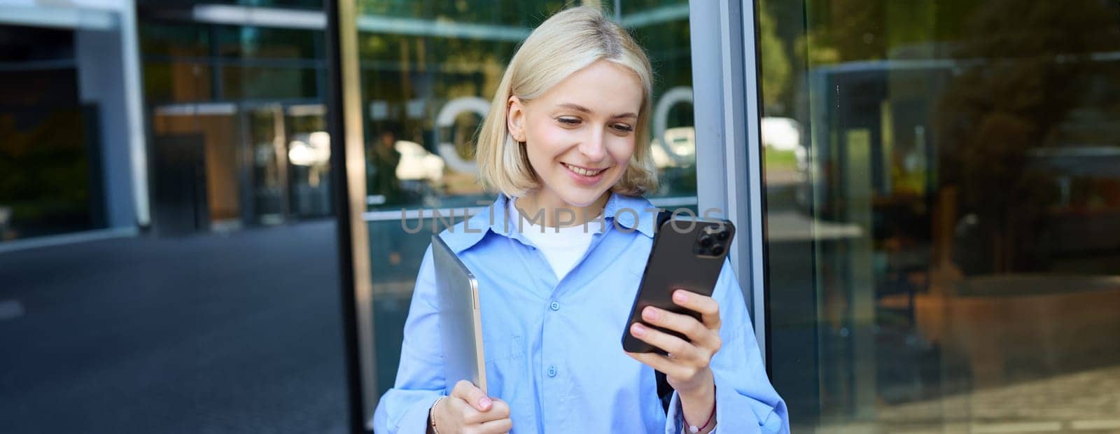 Image of young blond woman in blue shirt, holding laptop, waiting for someone near office building, using smartphone, mobile phone application by Benzoix