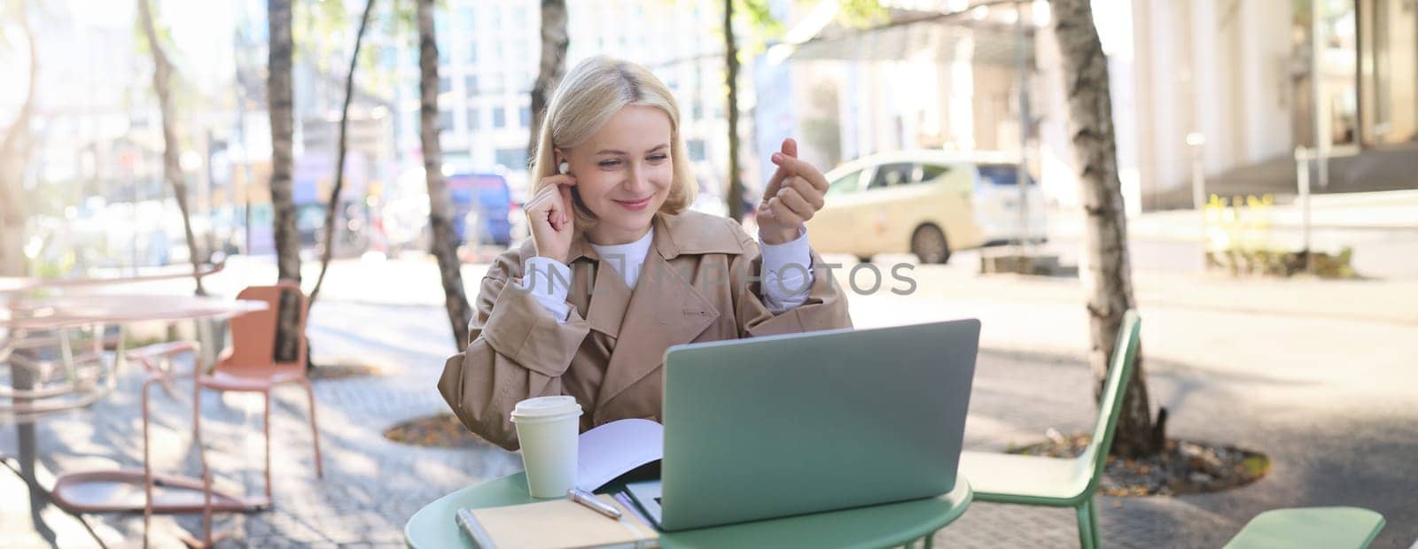 Cheerful young woman in trench coat with coffee, using wireless headphones and laptop, working remotely, connect to online class, video chatting by Benzoix